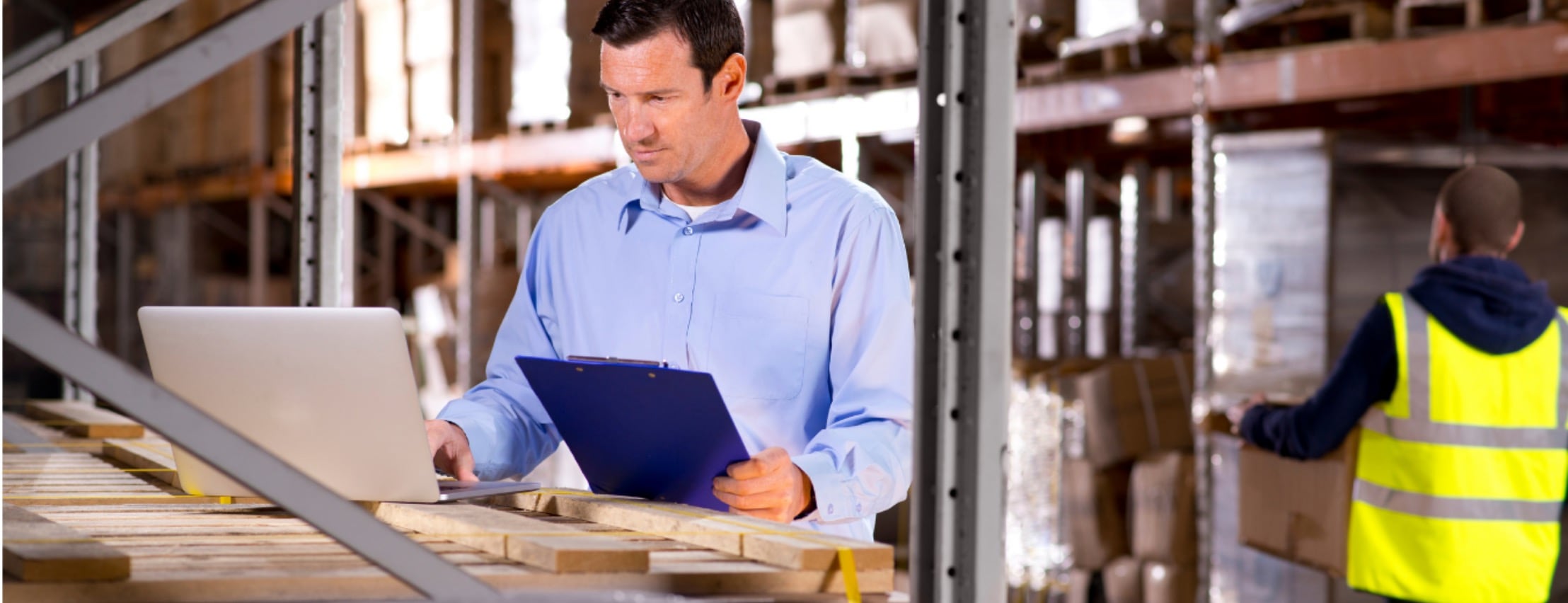 A man wearing a light blue button-up shirt types on a computer while holding a clipboard in a warehouse setting